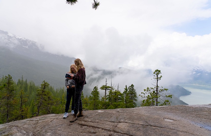 Two women referencing a guide book with view of Howe Sound through clouds