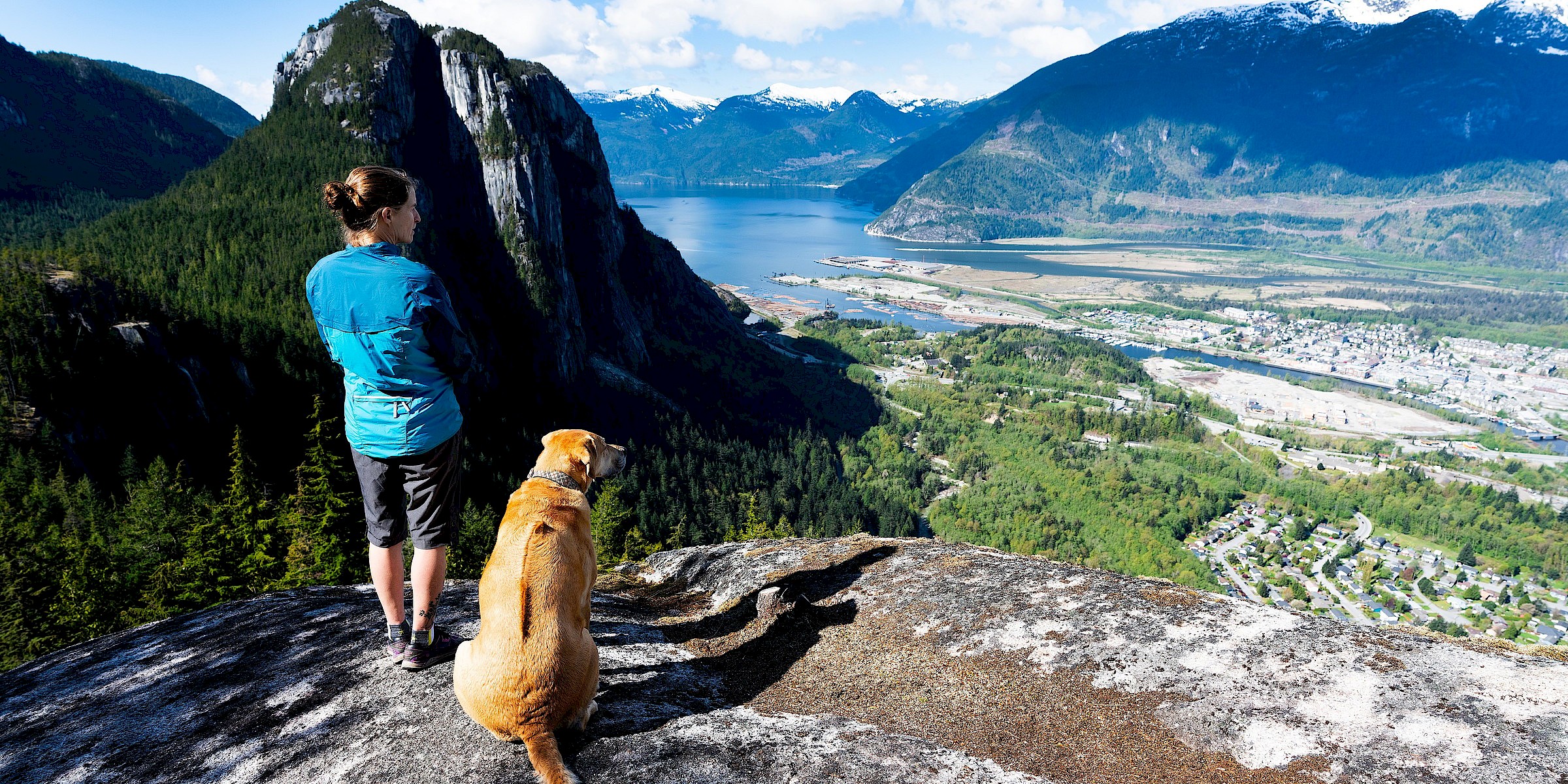 A hiker and her dog survey Squamish from the top of the Slhany